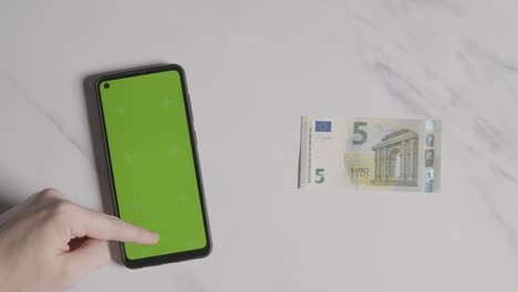 Overhead-Währungsaufnahme-Einer-5-Euro-Note-Neben-Einer-Person,-Die-Ein-Green-Screen-Handy-Verwendet