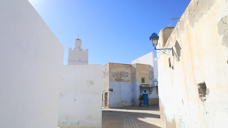 white buildings and a mosque in a narrow alley under the bright blue sky in an old tunisian city