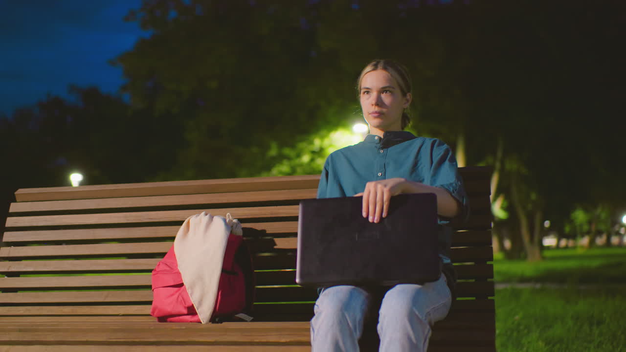 Young woman sits outdoors on bench at night with red bag nearby, opening laptop in a peaceful park setting, background includes trees, a light pole, greenery, and a cyclist riding behind