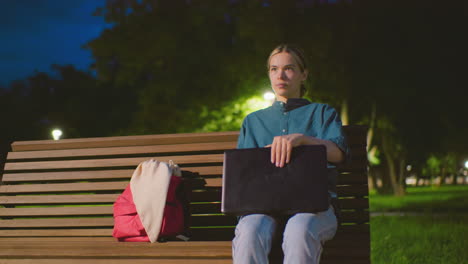 young woman sits outdoors on bench at night with red bag nearby, opening laptop in a peaceful park setting, background includes trees, a light pole, greenery, and a cyclist riding behind