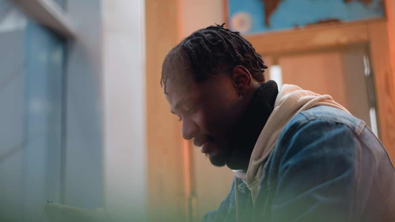 close up profile young black man wearing denim jacket hoodie reading tablet attentively in modern cafe setting with blurred green plant foreground warm ambient lighting wooden panel background