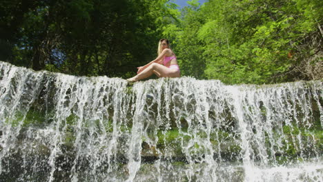 Confident-young-woman-in-bikini-sitting-and-posing-on-the-edge-of-a-small-waterfall,-wide-shot