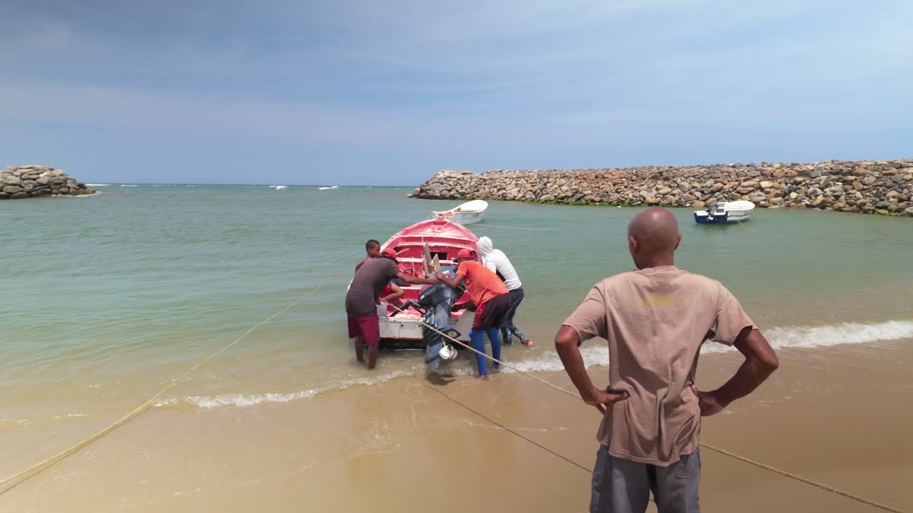 Two fishermen work together to adjust the engine of a boat on the shore. They are focused on their task while a third person observes from a distance.