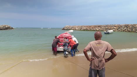 two fishermen work together to adjust the engine of a boat on the shore. they are focused on their task while a third person observes from a distance.