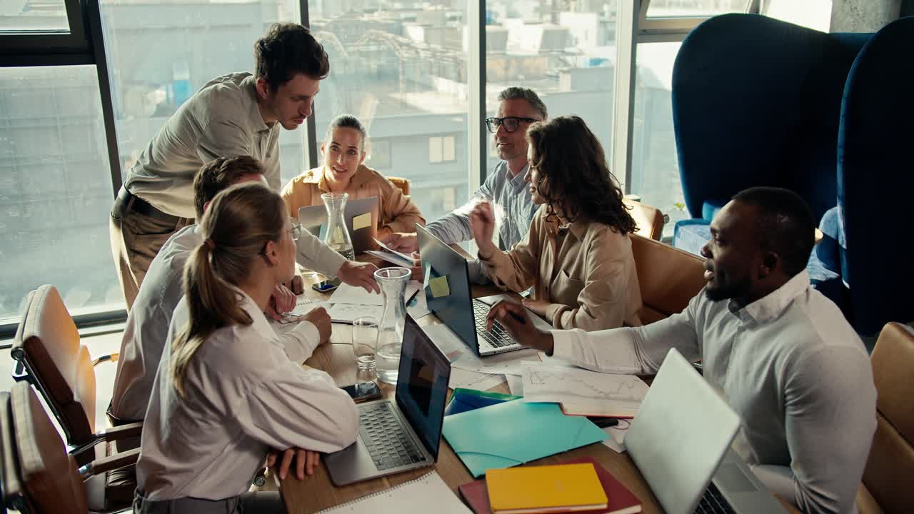A group of office workers sit at a table and show the manager guy in a white shirt their projects on paper. Team work at a table in an office with panoramic windows