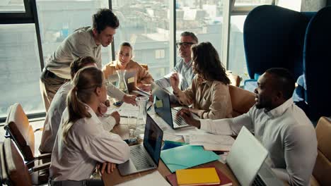 a group of office workers sit at a table and show the manager guy in a white shirt their projects on paper. team work at a table in an office with panoramic windows