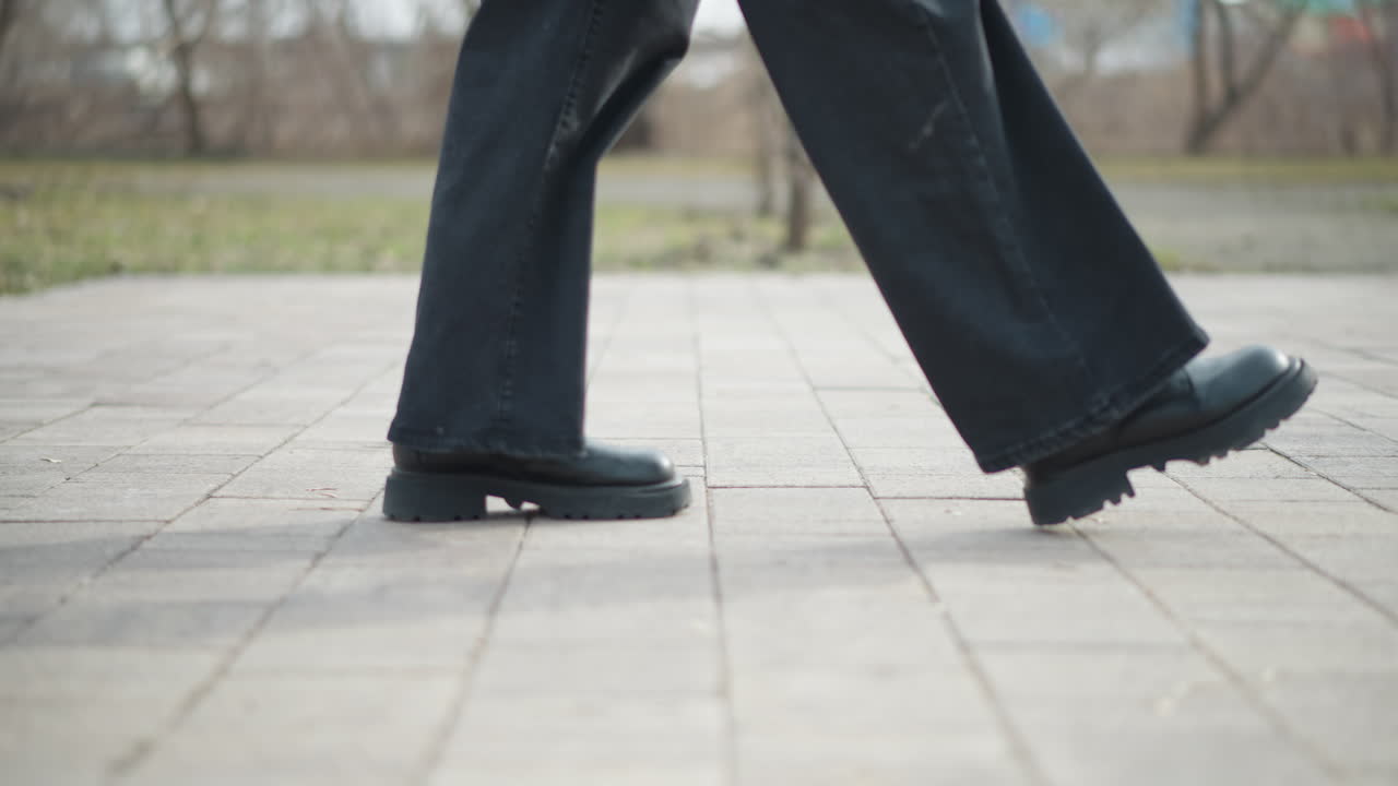 Side view close-up of person in black boots and dark jeans walking on paved surface in cold outdoor park, one foot slightly raised in mid-step, showcasing motion, balance, and calm winter atmosphere