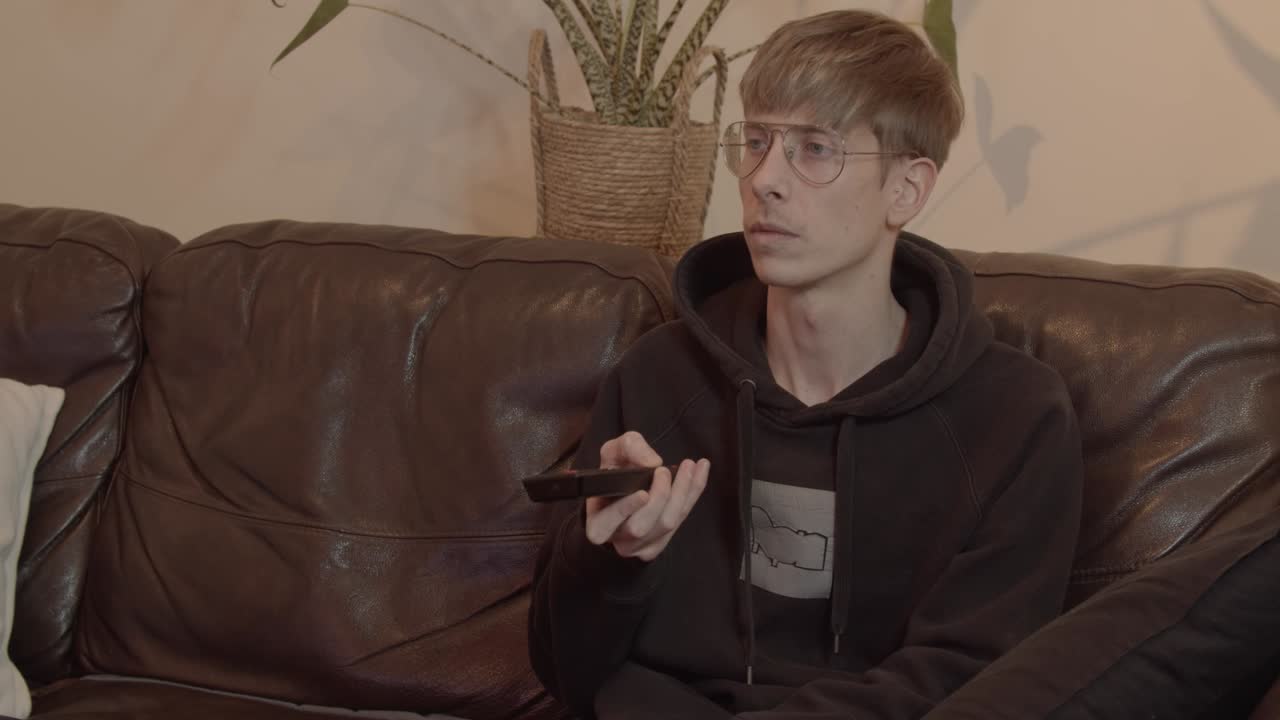 Young Man Watches TV and Uses Remote Control to Switch Channels