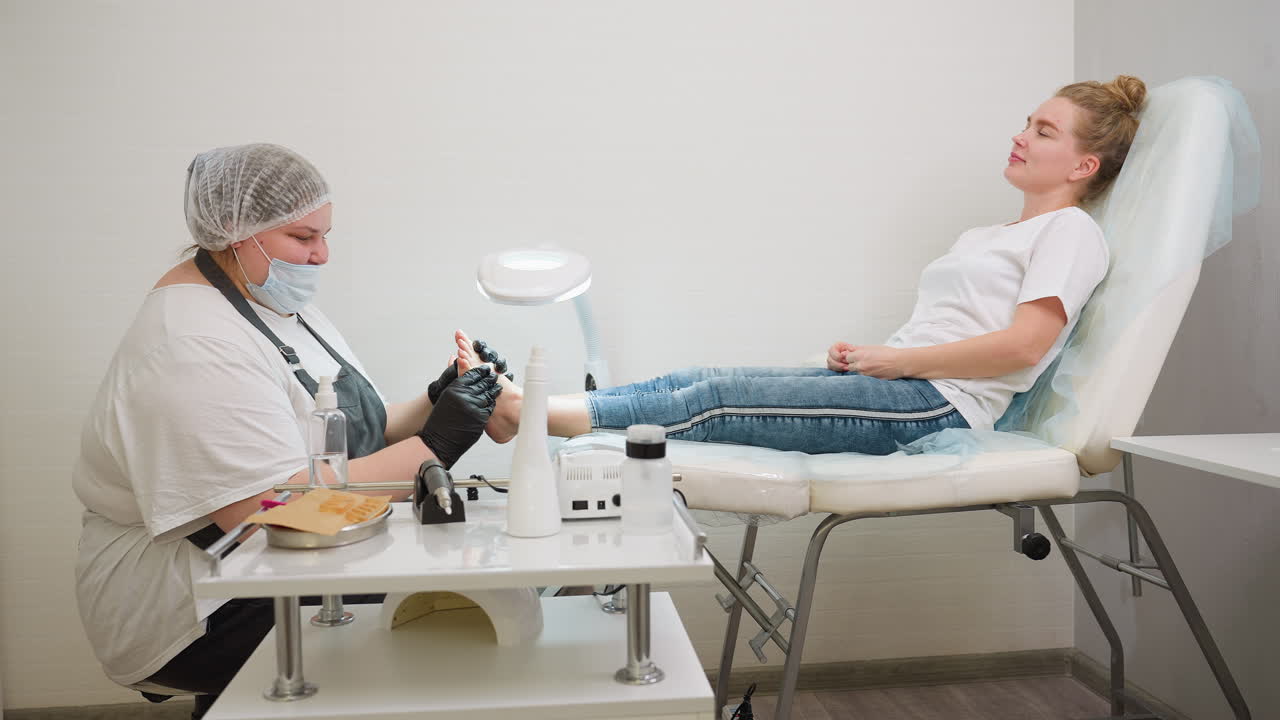 Nail technician wearing mask and gloves applies cream to client feet during pedicure session in bright salon setting, with professional tools arranged on nearby table for foot care procedure