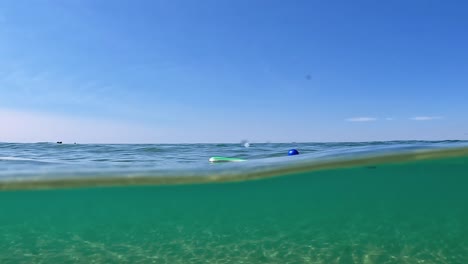 Ungewöhnliche-Geteilte-Unterwasseransicht-Von-Zwei-Verlorenen-Beach-Tennis-Paddeln-Und-Einem-Blauen-Ball,-Der-Auf-Der-Meeresoberfläche-Schwimmt