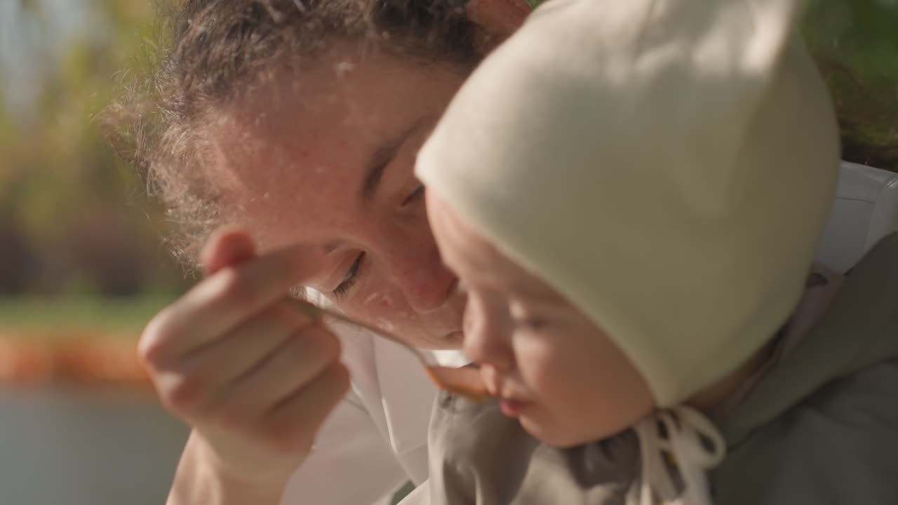 Madre che nutre il bambino con un cucchiaio in riva allo stagno, espressione giocosa e luce solare intensa, rami di salice e riflessi d'acqua lontani, intimità e fiducia familiare durante il pasto all'aperto.