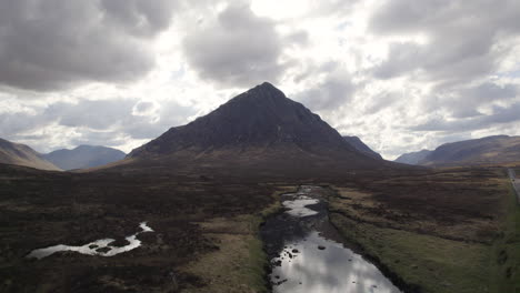 Volando-Bajo-Sobre-Un-Río-Poco-Profundo-Y-Un-Puente-Hacia-El-Pico-De-La-Montaña-Escocesa-Buachaille-En-Las-Hermosas-Tierras-Altas-De-Escocia