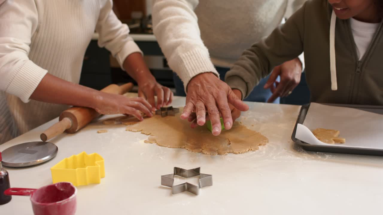 Una famiglia multiculturale che preme l'impasto dei biscotti sul bancone per tagliare delle forme usando uno stampino a stella, mentre il papà li guida.