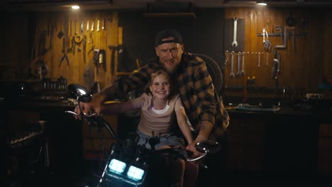 Portrait-of-a-happy-male-mechanic-with-a-beard-together-with-his-beloved-daughter-in-a-workshop-studio