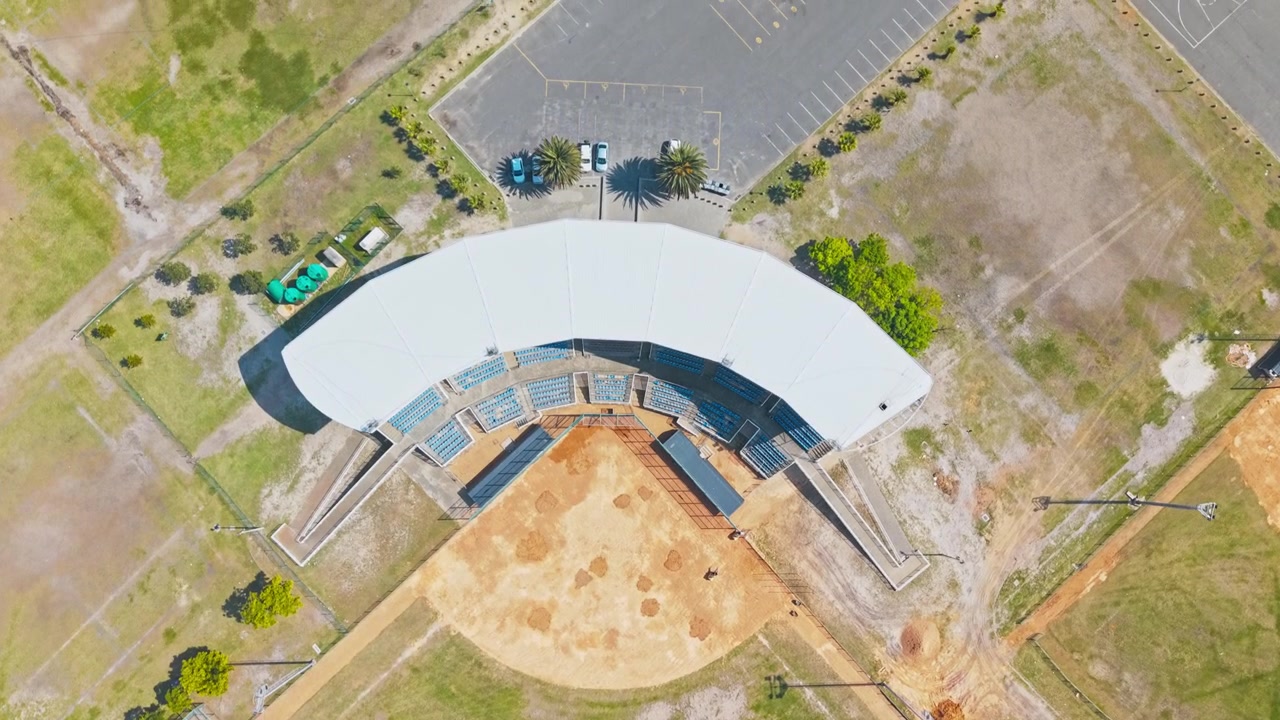 : A drone shot looks straight down on a baseball diamond, showing the sandy infield, curved outfield boundary, and surrounding green grass. Covered stadium seating with blue bleachers wraps.