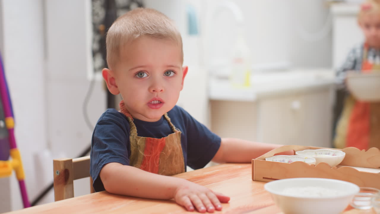 Bambino carino seduto a un tavolo che indossa un grembiule aspetta con innocenza, con le mani appoggiate sul legno, mentre una bambina si avvicina portando una ciotola durante una divertente attività in classe