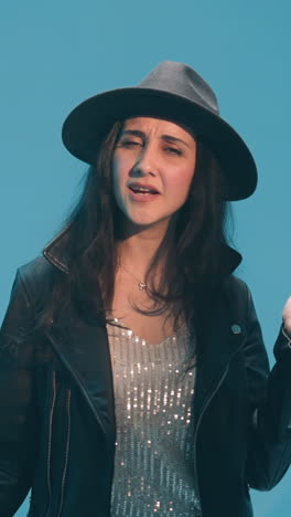 dama con sombrero negro y chaqueta de cuero cantando con gestos expresivos, sus manos y cuerpo en movimiento. un tecladista se ve en el fondo