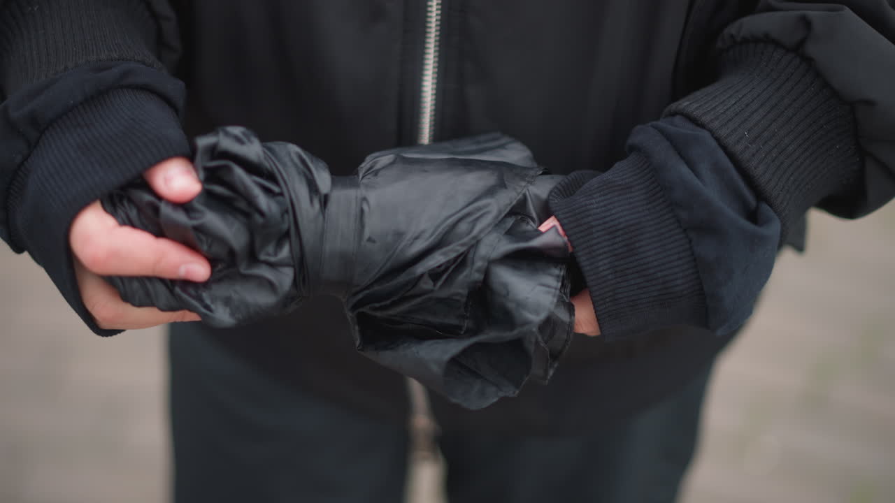Wet hands folding black umbrella on sidewalk, closeup of woman wearing torn knit gloves and zipped black jacket, raindrops clinging to fabric, reflections in puddles, moody urban commute atmosphere
