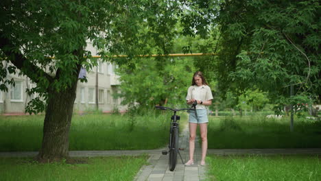 mujer con guantes negros se acerca a una bicicleta estacionada en un camino pavimentado, preparándose para usar guantes mientras usa un reloj inteligente en la mano derecha, el fondo incluye vegetación exuberante, árboles y edificio residencial