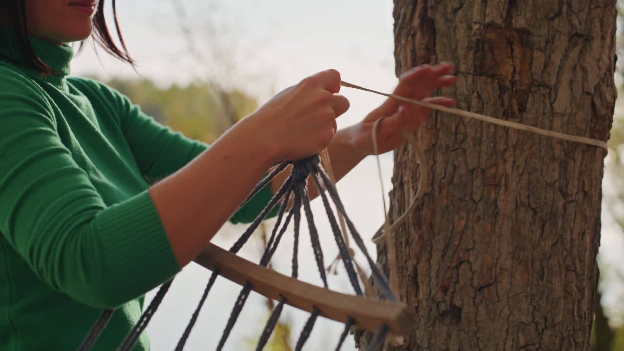 Woman tying hammock at lakeside, wearing green sweater and sunglasses, looping rope around tree trunk near calm water, slow focused movements, autumn light and serene solo camping preparation