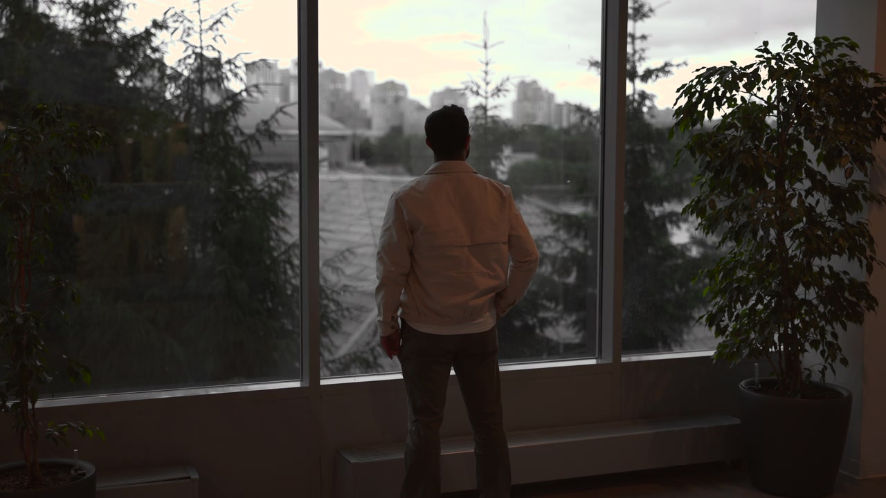 Single adult male seen from behind stands centered before floor-to-ceiling window, relaxed and still with hands at sides, overlooking city skyline through soft daylight inside quiet modern room