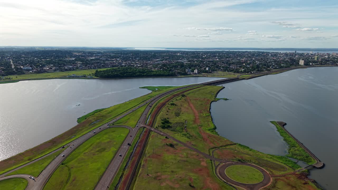 Forward aerial towards Posadas city via east road, with the Paraná River on the right, a river arm and bridge to the left, and lush vegetation including the botanical garden