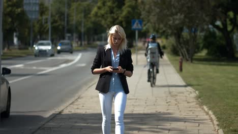 blonde beautiful young girl wearing white pants, blue dress shirt and blacktop walking down a busy city street with cars passing by on her phone checking social media and sending texts