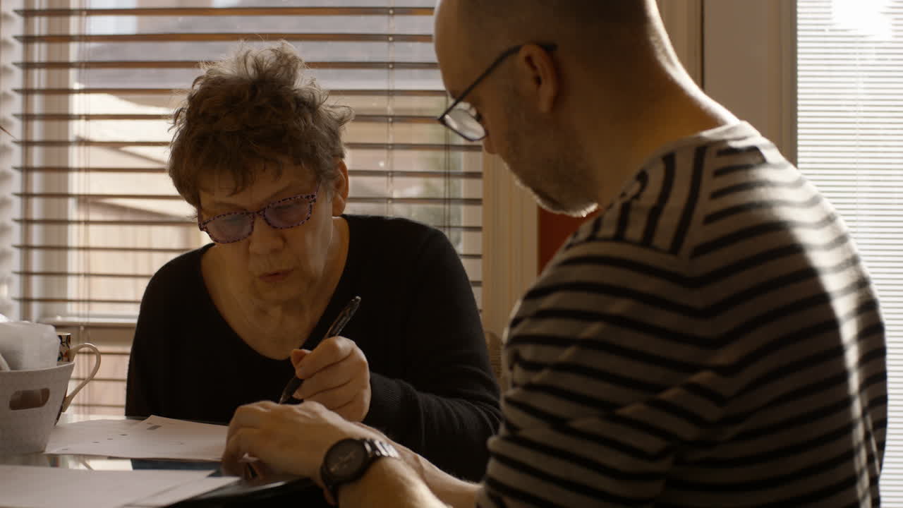 An elderly mother sits at a kitchen table with caregiver son. Mother reviews, document. Son hands her pen, to sign it. Both wear reading glasses. Warmly backlit by the sun through window. 24fps.