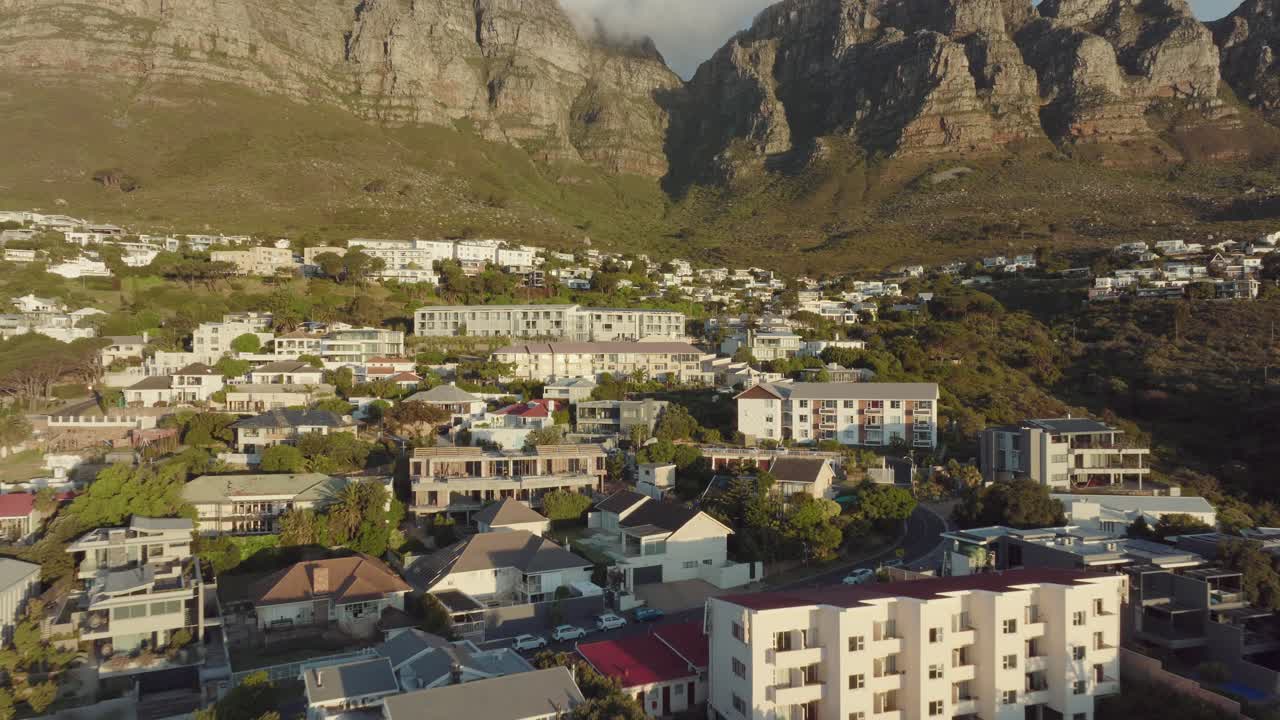 Drone flies fast backwards away from the houses standing on the hillside at Camps Bay beach in Cape Town South Africa - in the background Table Mountain rises illuminated by the sunset
