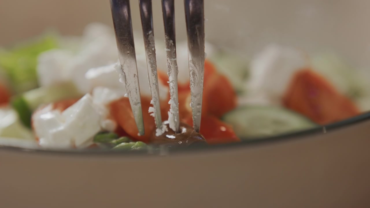 Detail vidličky míchající čerstvé salátové ingredience včetně rajčat, oliv, sýra feta a zeleniny. Akce se zaměřuje na interakci mezi náčiním a potravinami v měkkém osvětlení s rozmazaným.