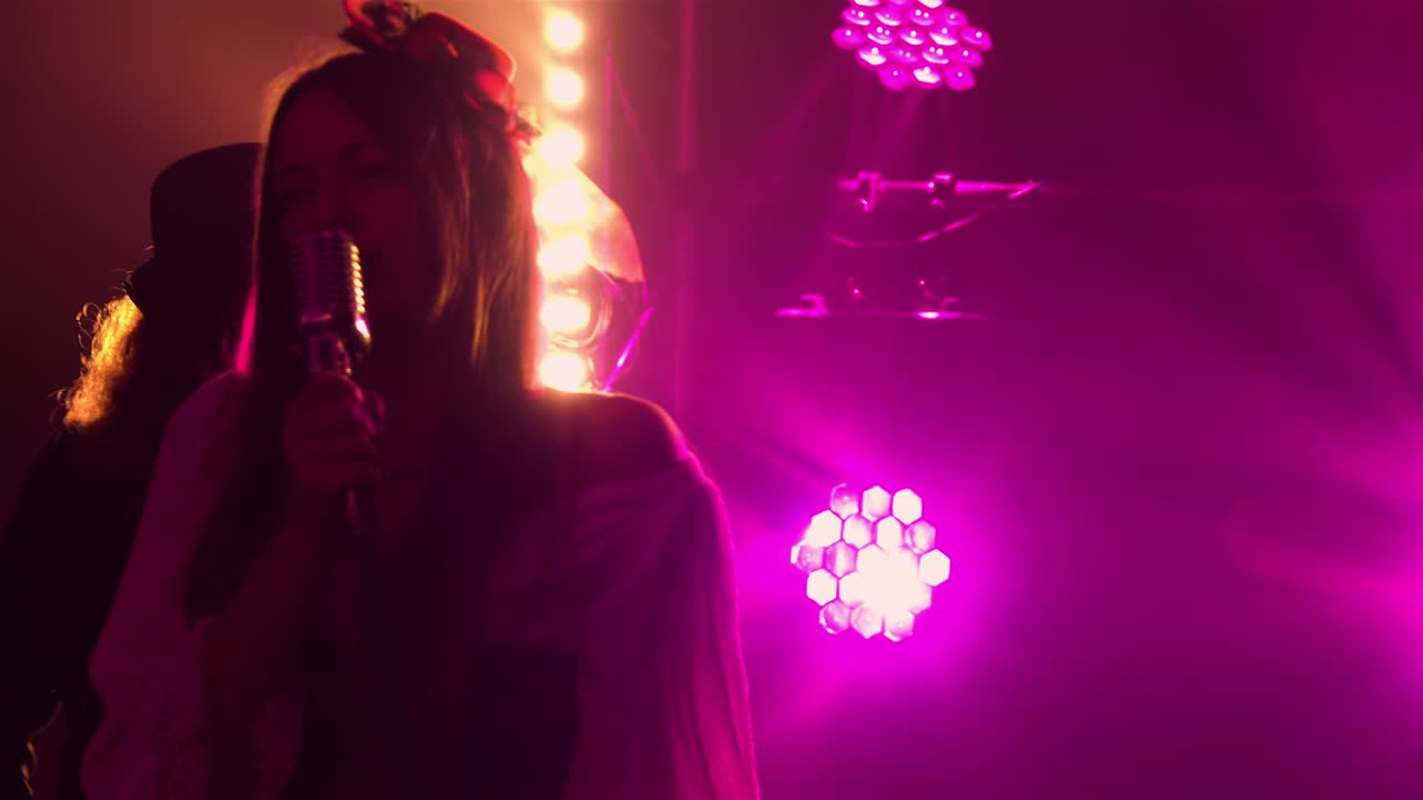 una elegante vocalista canta en el escenario en la oscuridad bajo la luz púrpura del estudio. en el fondo, un músico con un sombrero de estilo irlandés toca el violín. banda de rock, música enérgica, concierto en vivo, drive.