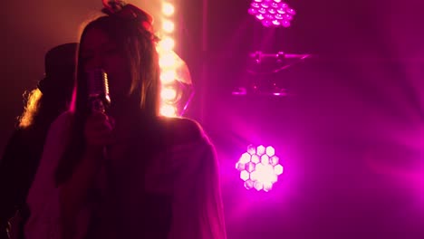 stylish female vocalist sings on stage in dark under purple studio light. in background, musician wearing an irish style hat plays the violin. rock band, energetic music, live concert, drive. close up