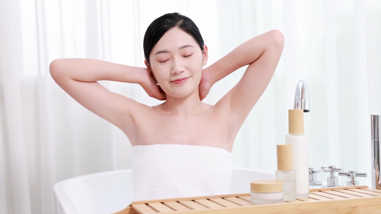 自宅で朝のスキンケアの前に、バスルームのバスタブでリラックスしながら腕を伸ばす、タオルに包まれた若く優雅な女性。スキンケア、セルフケア、ボディケア、スパのコンセプト
