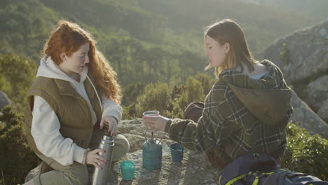 two young female hikers drinking hot tea from thermos while sitting at mountain cliff on a sunny autumn day