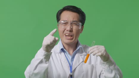 close up of angry asian man scientist looking at the orange liquid in the test tube and shouting to camera while standing on the green screen background in the laboratory