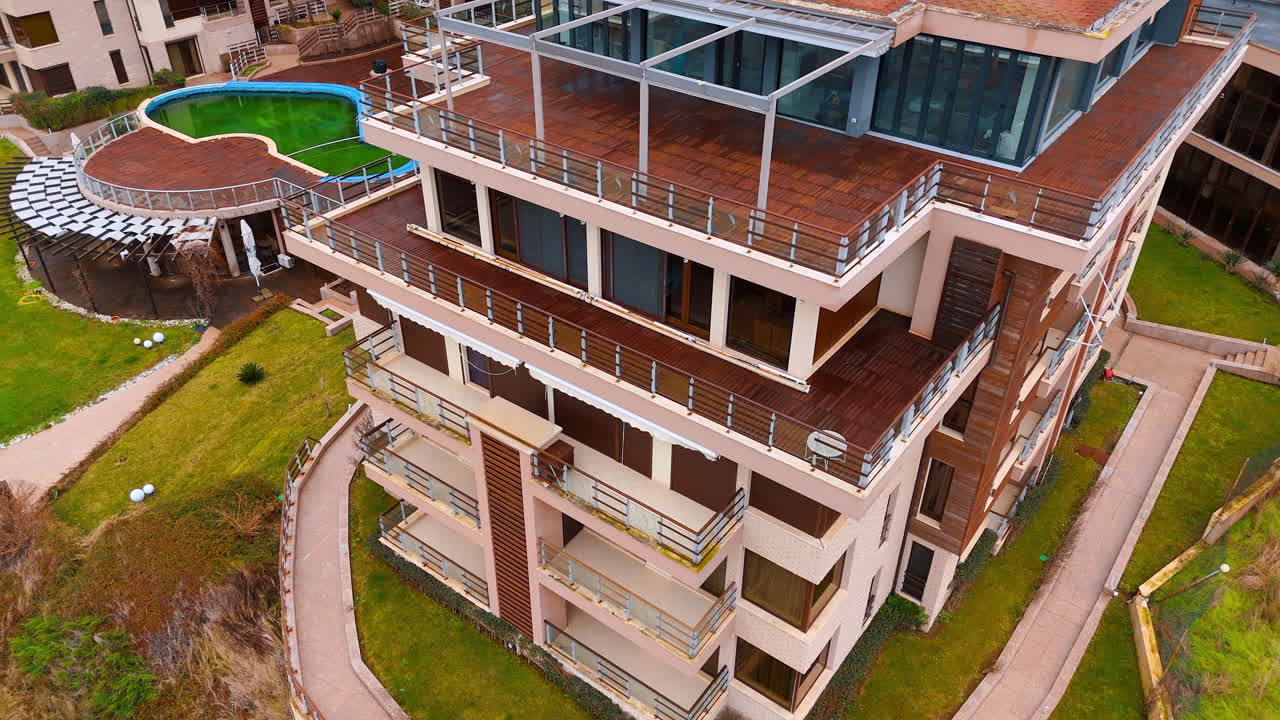 Vista aérea de edifício de apartamentos moderno com terraço e piscina. Edifício residencial de vários andares com terraço de madeira, piscina verde e caminhos de pedra vistos de cima.