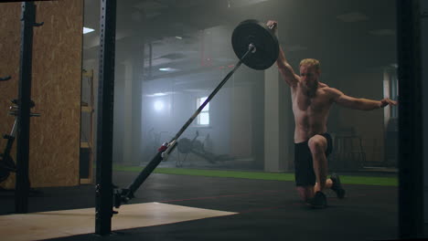 Un-Luchador-Masculino-Que-Hace-Ejercicio-En-El-Gimnasio-Empuja-Una-Barra-De-Minas-Terrestres-Con-La-Mano-Mientras-Está-De-Pie-Sobre-Una-Rodilla-En-Cámara-Lenta