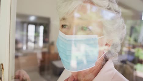 Sad-senior-caucasian-woman-wearing-face-mask-seen-through-window-in-slow-motion