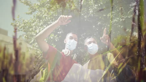 composite of happy biracial male friends in face masks reading map, and sunlight on trees