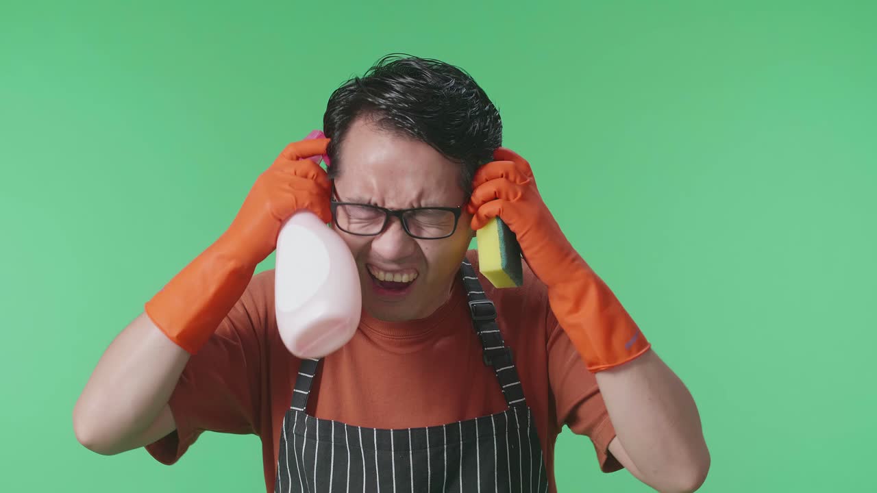 스프레이와 부 스폰지를 입은 아시안 남성 집주인이 녹색 스크린 스튜디오에서 두통을 고 있습니다.