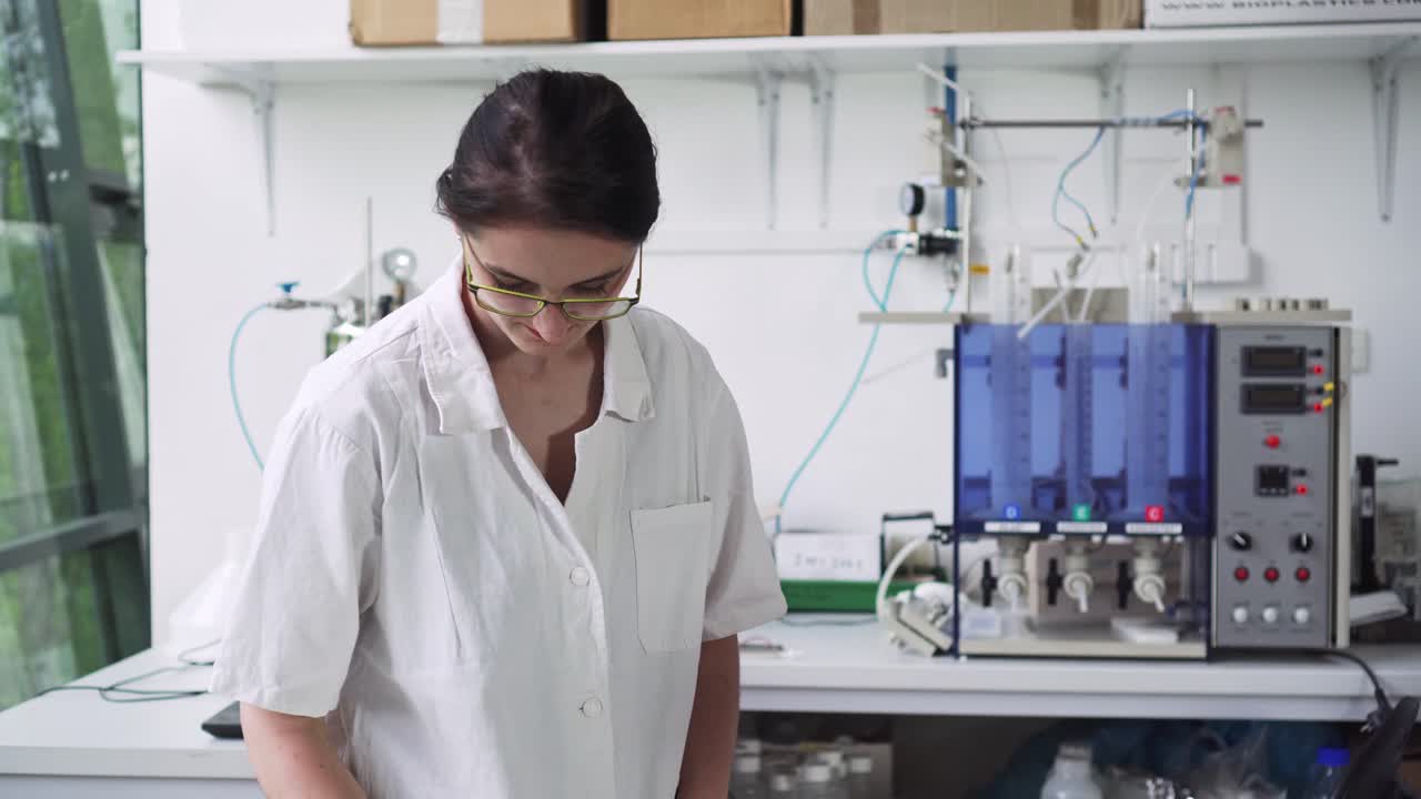 Pretty girl working in laboratory, marking test tube and using laptop, middle shot, front view