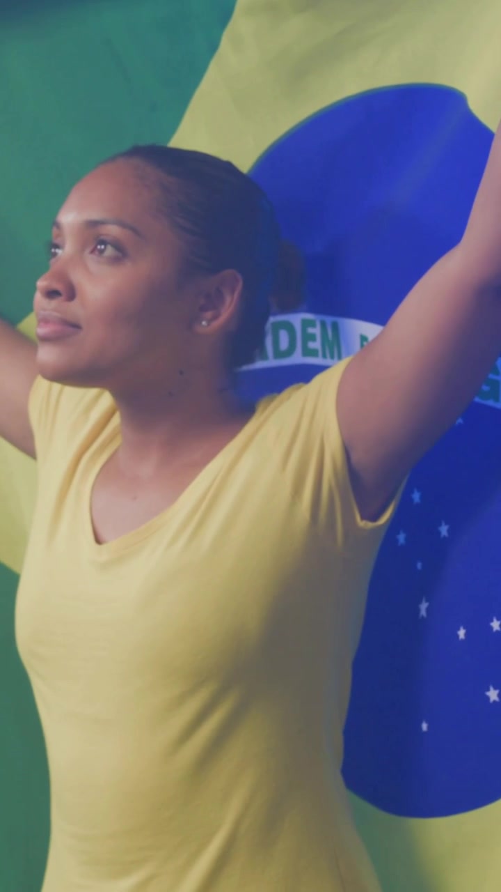 Vídeo Vertical: Mulher Entrando de Camisa Amarela Levantando e Abrindo a Bandeira Brasileira em Estúdio Mostrando o Emblema