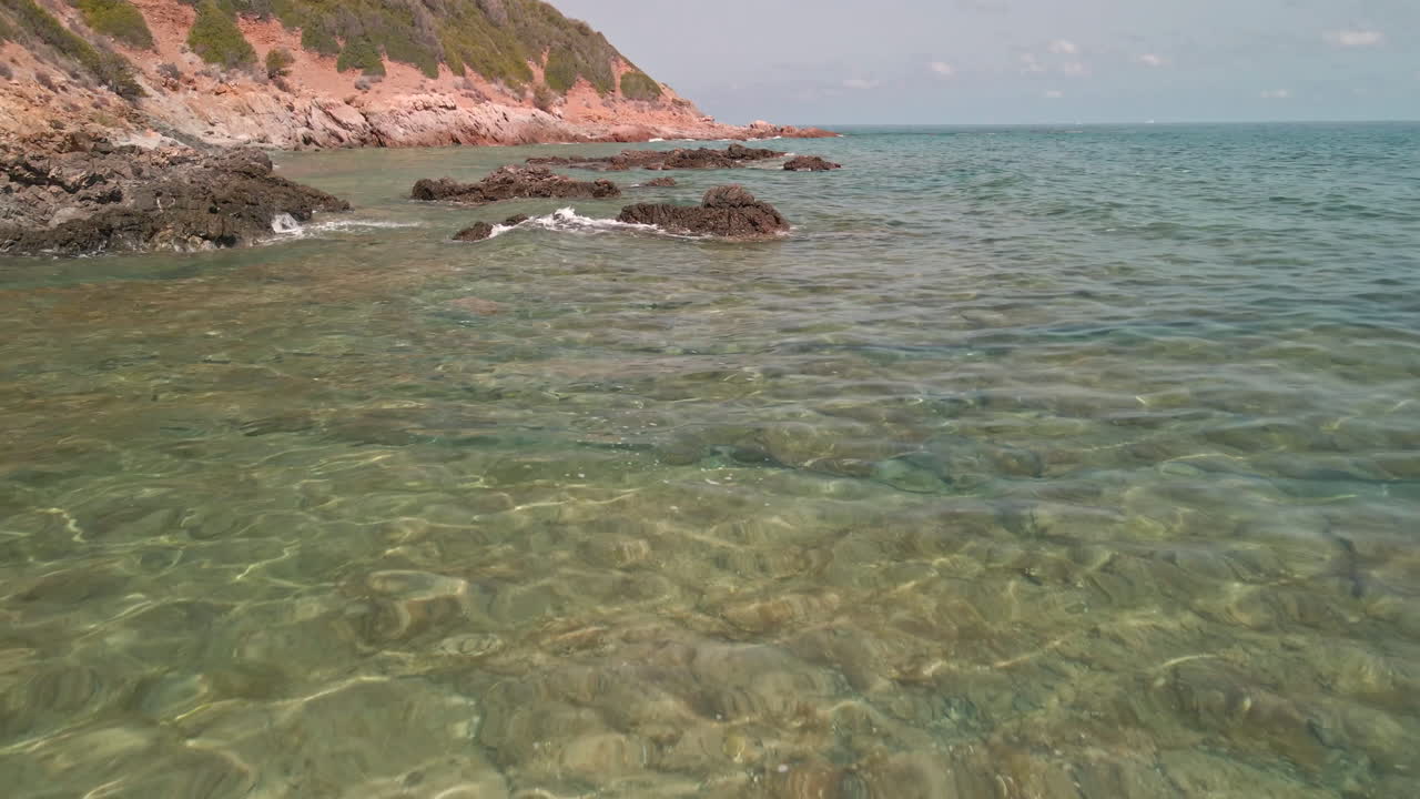 이탈리아 사르데냐에서 여름을 배경으로 푸른 초목이 있는 붉은 산에서 파도가 부서지는 바위 근처의 맑은 물가에서 영화 같은 드론 비행 - 공중 촬영