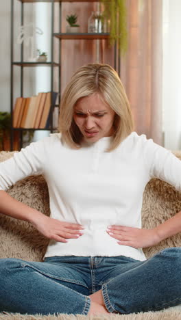 adult woman holding stomach at home expressing abdominal pain and discomfort while sitting on sofa