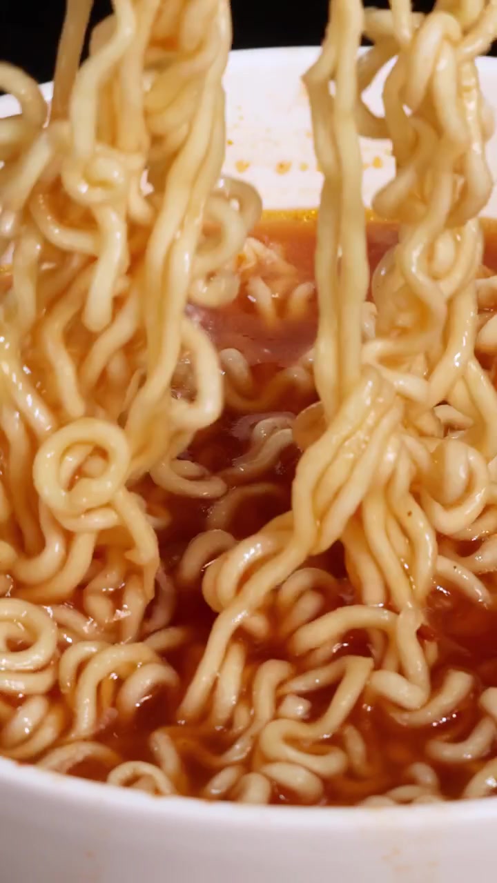 Forchetta che avvolge noodles istantanei piccanti in una ciotola bianca con brodo rosso sotto una luce intensa.
