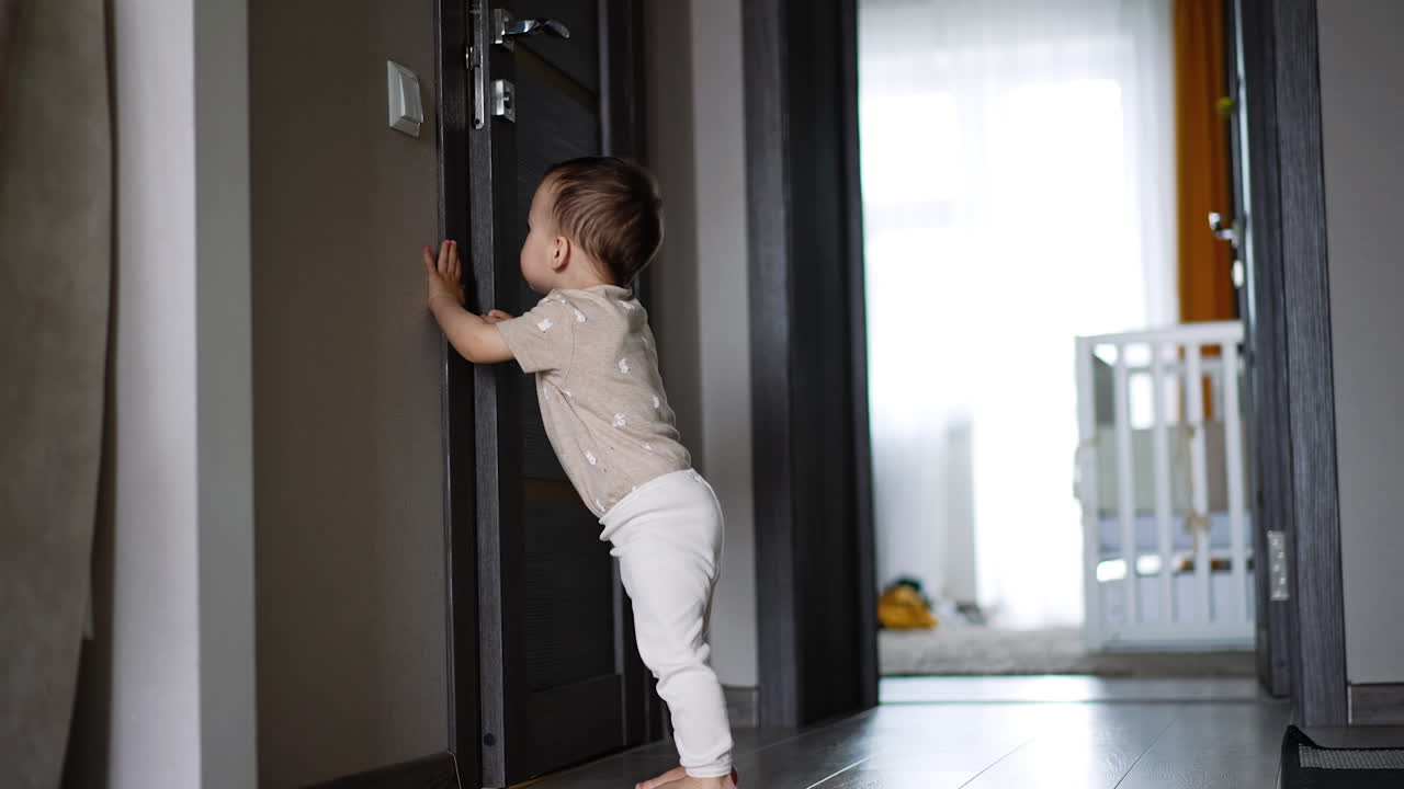 Cute toddler stands near the door. Kid pushes the door and looks how it opens ajar. Low angle view.