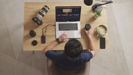 top view of asian male color grading drinking coffee while sitting in the workspace using a laptop next to the camera editing the video at home