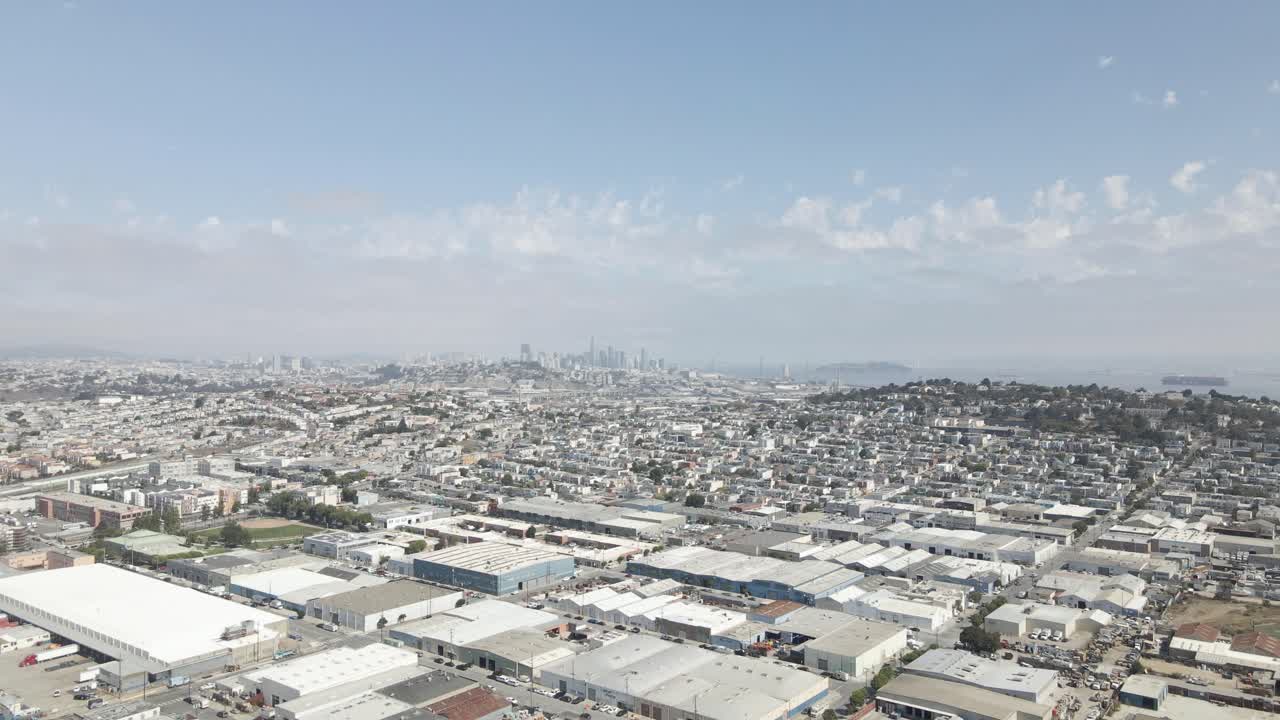 This pull out drone shot offers a sunny, scenic vista of San Francisco's Bayview neighborhood, commercial and residential areas, downtown skyline, and Yerba Buena Island, all vibrant bay activity