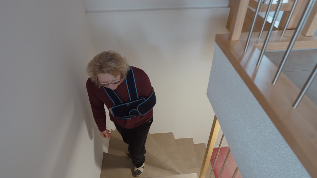 Authentic top view of an elderly woman with arm in sling climbing stairs at home. Concept of recovery, reduced mobility, aging, and everyday challenge