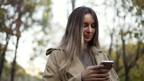 Lächelnde-Frau,-Die-Im-Herbstpark-Nachrichten-Liest-Oder-Vom-Telefon-Aus-Im-Internet-Surft